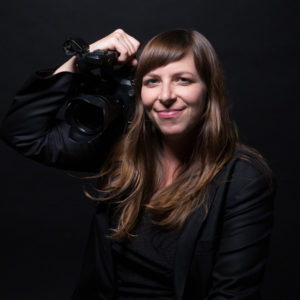 Portrait of Jen Gilomen, a white woman with long brown hair, holding her camera against a black background